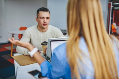 Ofis molasında elinde kahve tutan dalgın genç adam ve dikkatle dinleyen meslektaşı. Masaüstünde oturan yeni projeyle ilgili hipster duy ile iletişim kuran kadının arka planı.