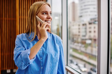 Akıllı telefon cihazından arkadaşınla iletişim kurarken camdan dışarı bakan mutlu, genç bir kadın. Ofisin içindeki modern selülomu çağırırken gülen olumlu hipster kızı.