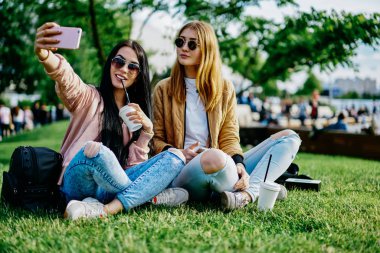 Genç bayan arkadaşlar parkta çimenlerde oturmuş akıllı telefon kamerasıyla selfie çekiyorlar. Hippi kızlar cep telefonlarıyla ortak fotoğraf çekiyorlar. Kampüste kahve molası veriyorlar.