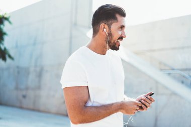 Beyaz tişörtlü beyaz adamın yan görüntüsü. Fotokopi alanı olan bir yere sahip. Elinde modern cep telefonu cihazları olan ve müzik listesi dinleyen ve teknoloji kavramını görmezden gelen beyaz bir adam.