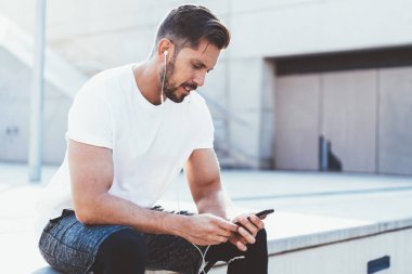 Spor kıyafetleri giymiş erkek sporcu dışarıda kardiyo eğitiminden sonra dinleniyor. Akıllı telefon zımbırtısıyla e-postaları kontrol ediyor ve koşmak için müzik listesi seçiyor. Elektronik kulaklıklı adam cep telefonu kullanıyor.