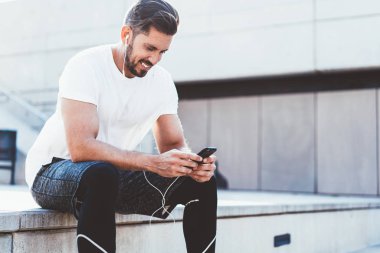 Aktif giyinmiş mutlu gülümseyen adam, dışarıda kardiyo eğitiminden sonra dinlenirken sağlıklı yaşam tarzı hakkında kitap okurken kulaklıklı neşeli bir adam, uygulamadan müzik dinlemekten keyif alıyor.