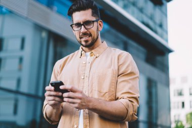 Modern iletişim teknolojisi olan yakışıklı beyaz adamın portresi. Şehir ortamında dikilip poz veren, optik gözlüklü akıllı telefon kullanıcısı kameraya bakıyor.