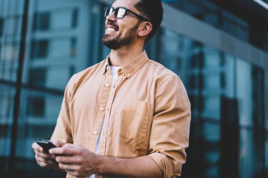 Dijital teknolojinin neşeli kullanıcıları ellerinde cep telefonuyla şehir ortamında dikiliyor ve gülümsüyor, sıradan giyinen mutlu beyaz adam akıllı telefon aleti tutuyor ve eğleniyor.