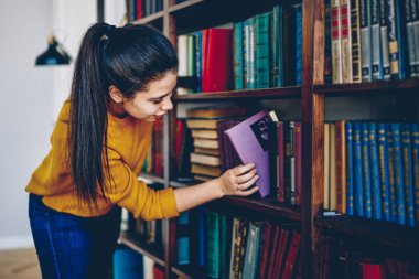 Young caucasian woman student searching literature for reading and education in university library, positive hipster girl looking for interesting novel choosing books on shelves in bookstore clipart