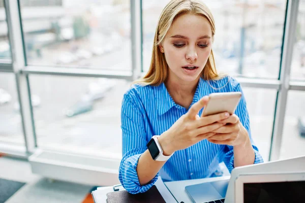 Young woman from millennial generation typing text messaging on modern smartphone and chatting online in social networks using wireless internet connection in office.HIpster blogger checking email