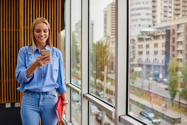 Cheerful female blogger reading funny notification in social networks on modern smartphone device using free 4G internet.Positive young woman chatting online on cellular standing in office interior