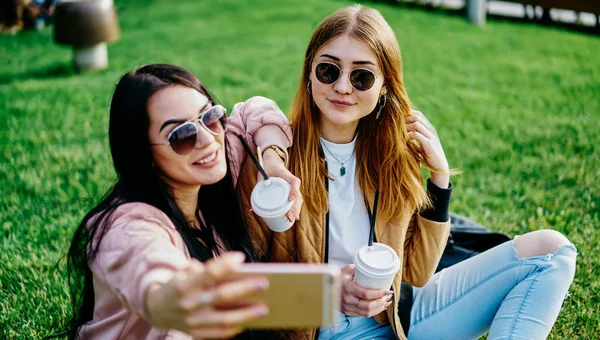 ブルーレットとブロンドの女性ブロガーは スマートフォンのカメラは公園で夏時間を過ごすためにポーズ ヒップスターの女の子の友人は一緒にソーシャルネットワークのプロフィールで共有のための写真を撮るコーヒーブレイクを楽しんで — ストック写真