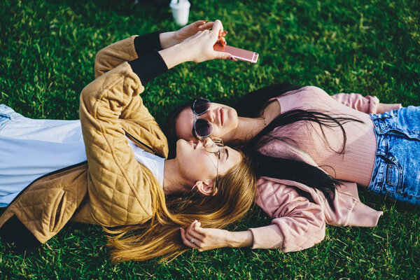 Young female friends lying on green  grass together in trendy sunglasses using smartphone camera for selfie, pretty hipster girls in stylish wear taking picture on mobile phone resting in park