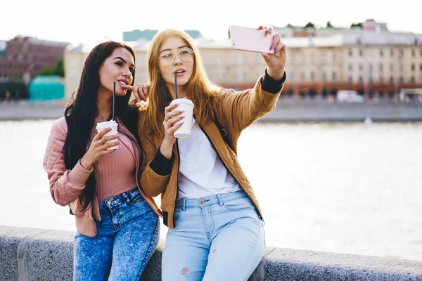 Attractive young women travelers making photo on smartphone camera drinking coffee to go on urban settings, female friends posing for selfie on mobile phone spending free time on street together