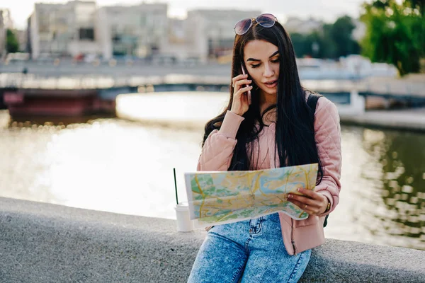 Young brunette woman having conversation in roaming calling to operator for booking taxi during trip in town, female traveler talking with friend  for arrange meeting checking route on map outdoors