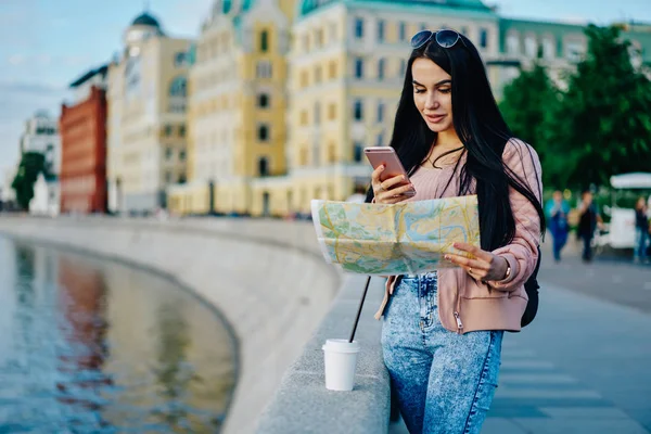 Positive female traveler using mobile phone app for checking location while holding map, caucasian hipster girl using internet in roaming holding smartphone while planning route of showplaces