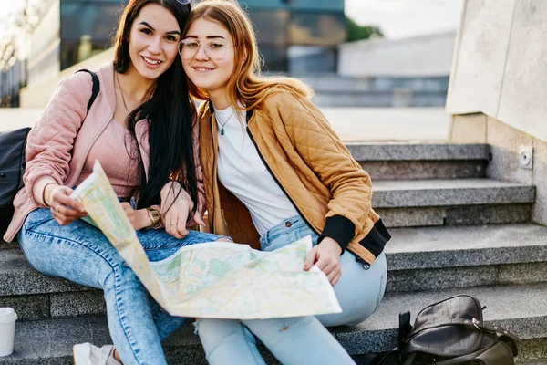 休暇に満足した陽気な女性の親友の肖像画一緒に都市の設定に座って 幸せな原因アジアの女性観光ツアーへのルートを計画するための地図を使用してカメラを見て旅行 — ストック写真