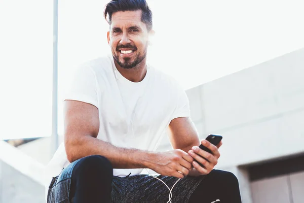 Portrait of cheerful male jogger sitting outdoors with cellular phone in hand and smiling at camera during time for training in city, happy man in headphones enjoying music sounds from website