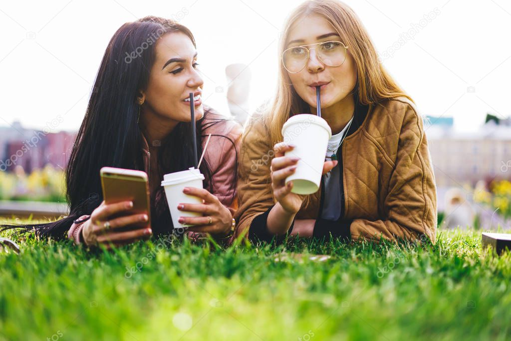 Young blonde and brunette women drinking beverage and lying on green grass having friendly communication, positive hipster girls talking and recreating during summer free time with coffee to go