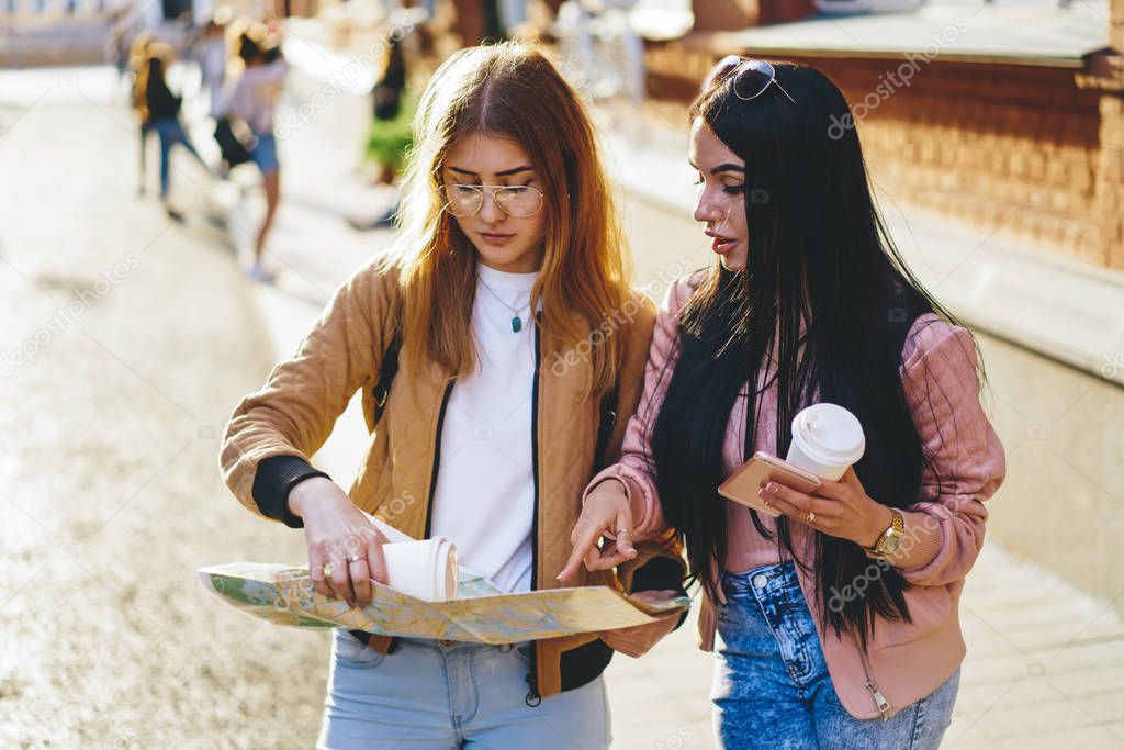 Brunette and blonde female tourist using map for strolling in city and getting to showplaces, women best friend talking to each other choosing route for walking during spring vacation together