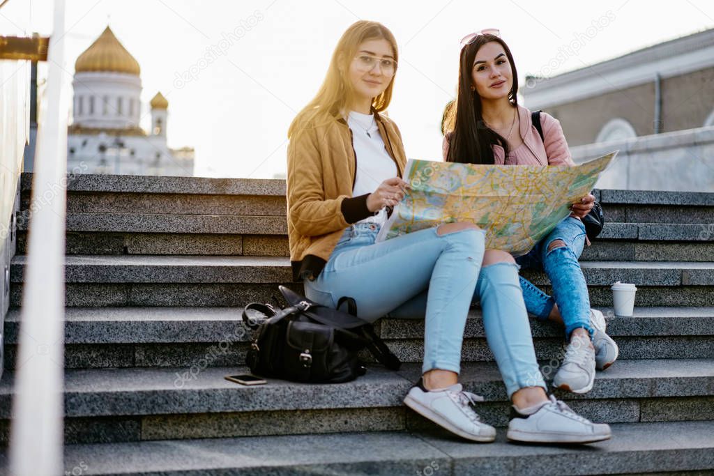 Portrait of cheerful brunette and blonde female best friends spending spring vacation together in city, young female travelers sitting with map on stairs for planning route looking at camera