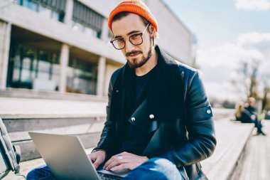 Modern netbook 'la açık havada oturan yetenekli bir adamın portresi, görüş düzeltmesi için gözlüklü beyaz tenli hippi şehir merkezindeki uzaktan çalışma sırasında kameraya bakıyor.
