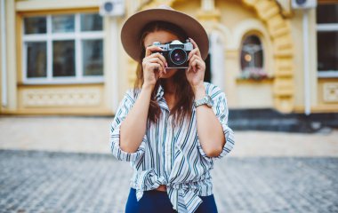 Sokakta fotoğraf çekmek için klasik kamera kullanan modaya uygun şapka ve giysili genç bir kadın, açık havada kentsel ortamların resmini yapan profesyonel bir kadın fotoğrafçı.