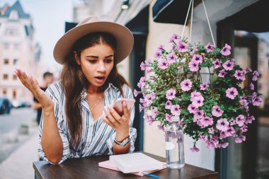 Moda şapkalı duygusal hippi kız caddedeki kafede cep telefonuyla kötü internet bağlantısını karıştırıyor, genç kadın akıllı telefondan bankacılık servisinden gelen uyarıları kontrol ederken merak ediyor.