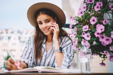 Şapkalı çekici beyaz kadın cep telefonuyla konuşurken kameraya bakıyor, mutlu hipster kız portresi akıllı telefon konuşması yapıyor not defterine bilgi yazıyor.