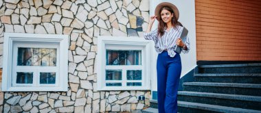 Moda kıyafetleri içinde ve sokakta yürürken şapka takan, şık giyinmiş, şık giyinmiş, neşe dolu beyaz kız elinde kitap tutarak sokakta vakit geçiriyor.