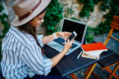 Şapkalı olumlu hipster kız cep telefonu ve 4G bağlantısı kullanarak sosyal ağlarda yeni beslemeleri kontrol ediyor, kadın blogcu medyayı ve multimedya içeriğini paylaşıyor sokakta dizüstü bilgisayarla kafede oturuyor