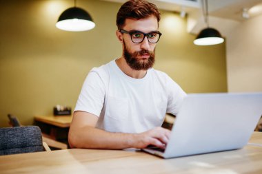 Gözlüklü ciddi bir erkek blogcu dizüstü bilgisayarda yayınlanması için makale yazıyor, yetenekli hipster adam internetteki internet dersini birlikte çalışma odasında izlemeye odaklanmış.