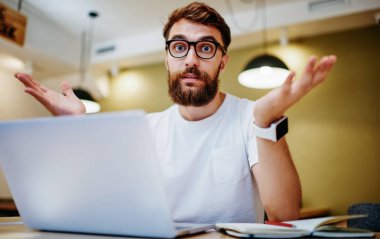 Kafası karışmış gözlüklü beyaz erkek öğrencinin portresi dizüstü bilgisayardaki yazılım arızasıyla karışmış, kameraya bakan endişeli hippi adam netbook üzerinden omuz silkiyor.