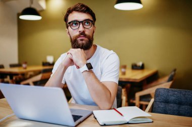 Gözlüklü, düşünceli, beyaz bir erkek birlikte çalışma alanındaki netbook 'ta uzaktan kumandayla çalışarak yayınlama fikirleri üzerinde kafa yoruyor. Düşünceli hipster adam, kafeteryadaki çevrimiçi projeye yoğunlaştı.