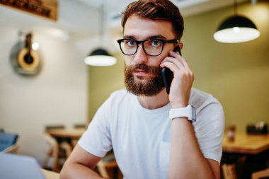 Ciddi bir beyaz sakallı adamın portresi cep telefonuyla bankacılığı arıyor içeride oturuyor, görüş düzeltmesi için gözlüklü genç bir hippi kameraya bakıyor telefonda konuşuyor.