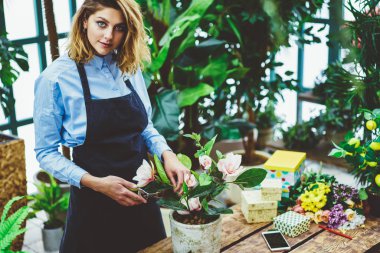 Çiçek hobisi için harcadığı süre boyunca çiçekçilikte bahçeyle uğraşan pozitif botanikçinin yarım boy portresi. Profesyonel önlüklü başarılı bir kadın kendi konservatuarında dikilirken kameraya bakıyor.