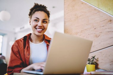 Neşeli hippi kız modern netbook ile birlikte çalışma alanında oturuyor ve komik mesajlar alıyor, olumlu siyah kadın kafasını çeviriyor ve kafenin içinde öğrenme sırasında gülümsüyor.