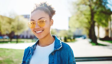 Güneş gözlüklü pozitifAfrikalı bir kadın şehir ortamının yanında durup bahar havasının tadını çıkarırken kameraya bakıyor. Şehre doğru yürürken giyinmiş mutlu hippi bir kızın portresini yakından çekiyor.