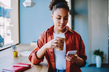 Kafein içeceklerinin tadını çıkarırken akıllı telefon cihazıyla mesaj gönderen çekici, koyu tenli hippi kız, elinde kahve ile elinde kağıt bardak tutan Afrikalı kadın ve web sitesinde politik haberler okuyor.