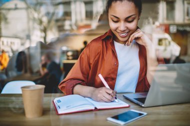 Üniversite kafeteryasında öğrenim ve sınav hazırlığı, bilgi ve otodiktat kavramı sırasında eğitim için ders kitabında web sitesinden bilgi yazan koyu tenli bir kız öğrenci