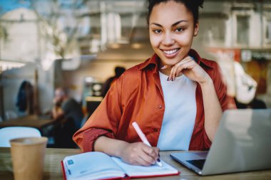 Kameraya bakıp öğrenmenin tadını çıkaran başarılı bir kız öğrenci. Dizüstü bilgisayarın önünde otururken gülümseyen Afrikalı bir kadının portresi. Not defterinde ay planlaması ve Netbook 'ta organizatör kullanması.