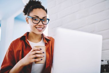 Optik gözlüklü, serbest çalışan, elinde yemek fincanı tutan ve reklam alanında kahve molası veren yetenekli bir kadın portresi. Kafeteryada dizüstü bilgisayarlı mutlu koyu tenli hippi kız.