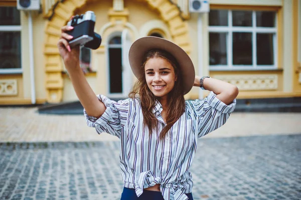 Cheerful caucasian woman in trendy hat making photo of herself  using vintage camera  on street, beautiful teen hipster  posing for picture dressed in stylish outfit spending free time in town