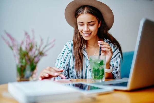 現代的な技術とWifiを使用してカクテルとカフェで休憩中に休憩中の流行の帽子で笑顔ヒップスターの女の子 陽気な魅力的な女性フリーランサーは ネットワーク内のガジェットブログを介してリモートで作業 — ストック写真