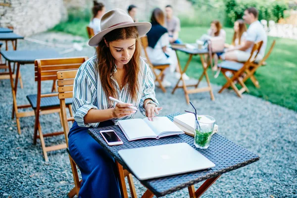 集中ブルネットヒップスター女の子で帽子注目情報でノートブック作りにリストを行うにはながら 屋外のカフェでテーブルで休んで 集中的な白人女性ブロガーノートブックに記事を書く — ストック写真