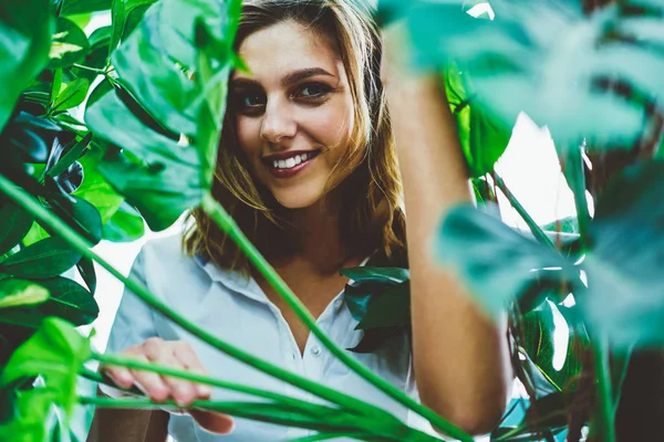 緑の葉の近くに立つ陽気で興奮した白人女性の花屋の肖像画を閉じ 植物園からの休憩中に顔にかわいい笑顔でカメラを見る植物園 — ストック写真