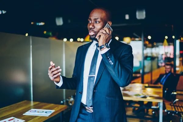 Dark skinned male entrepreneur dressed in suit discussing business plan during mobile conversation on smartphone device.African American proud ceo calling on cellular device while working in office
