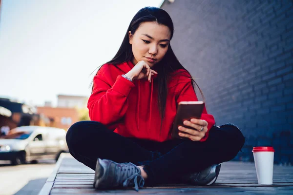 Young japanese woman sitting outdoors with mobile phone connected to 4G internet in roaming, teenager hipster girl sending text messages via smartphone chatting with followers in social networks