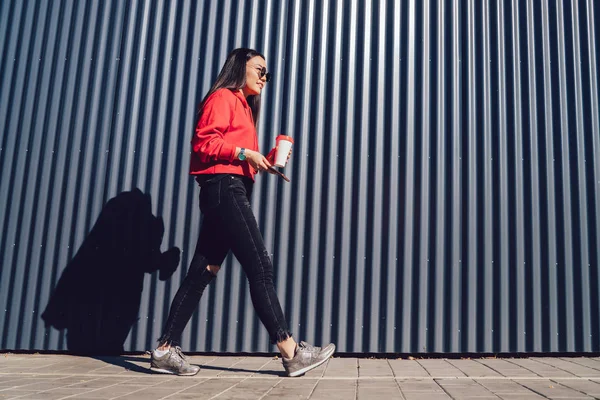 Attractive chinese hipster girl with coffee to go and smartphone device in hands strolling to meeting with friend on urban setting, beautiful millennial woman walking with takeaway paper cup