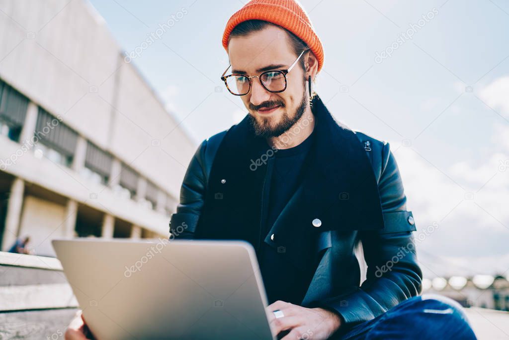 Successful male freelancer in optical eyewear for vision correction searching information for creating project via laptop application, positive hipster guy in stylish wear reading news on website