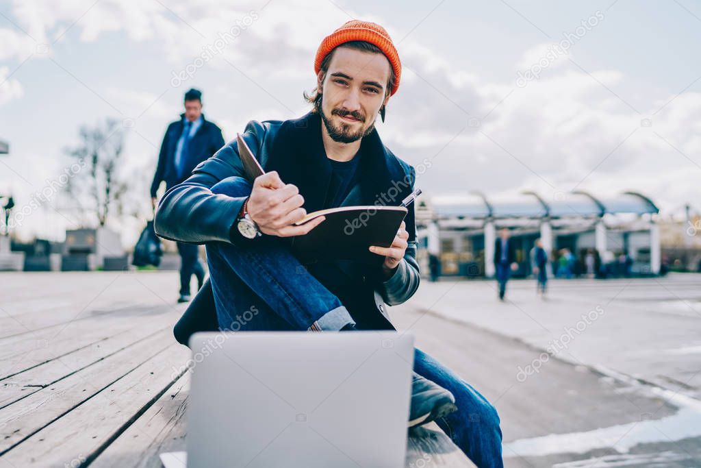 Portrait of positive male student in stylish outfit looking at camera and feeling good from spending time for autodidact and e learning, handsome hipster guy holding sketchbook for designing in hands