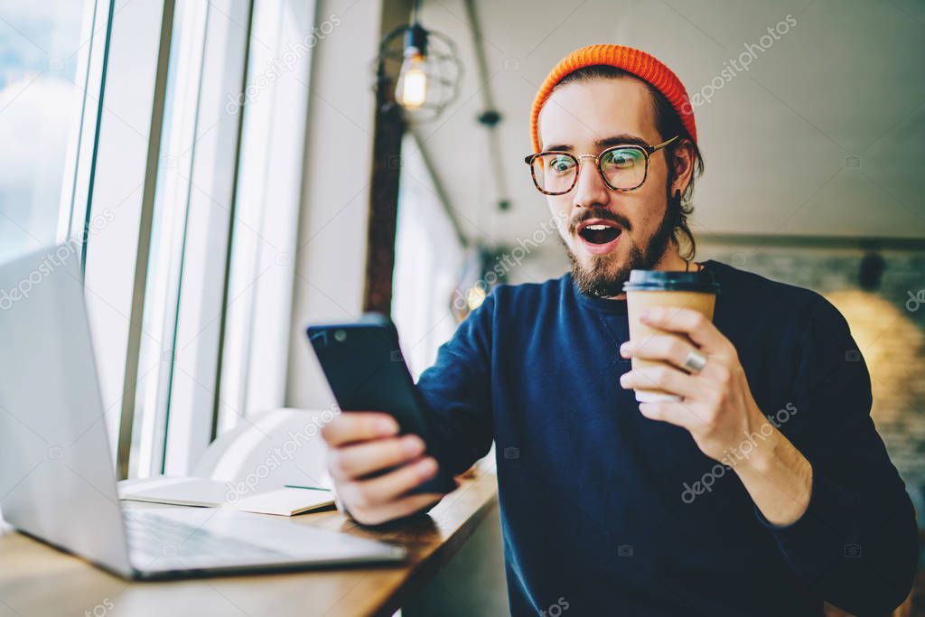 Amazed hipster guy watching shocking content news via application on cellphone and feeling wonder. Surprised male blogger reading received message. Millennial man phoning with shopping promotions