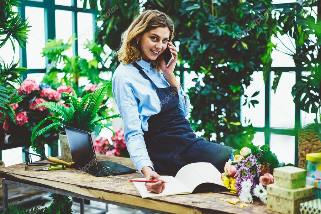 Portrait of happy female florist spending time at own flowers shop and calling to provider for consultancy about fresh blossom, successful owner of small business talking with colleagues via cellular