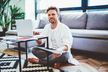 Uzaktan kumandayla yetinen beyaz erkek bir ressam apartman dairesinin modern iç odasında oturuyor, netbook ve evdeki kablosuz interneti kullanarak kameraya bakan mutlu genç hippi.
